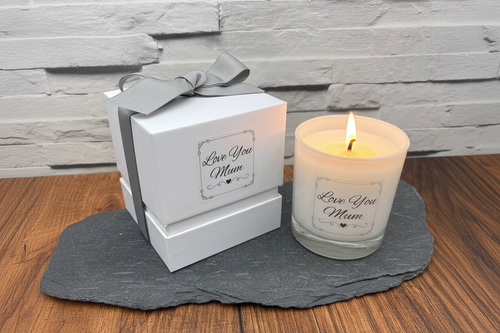 Candle with 'Love You Mum' message next to a gift box on a wooden surface.